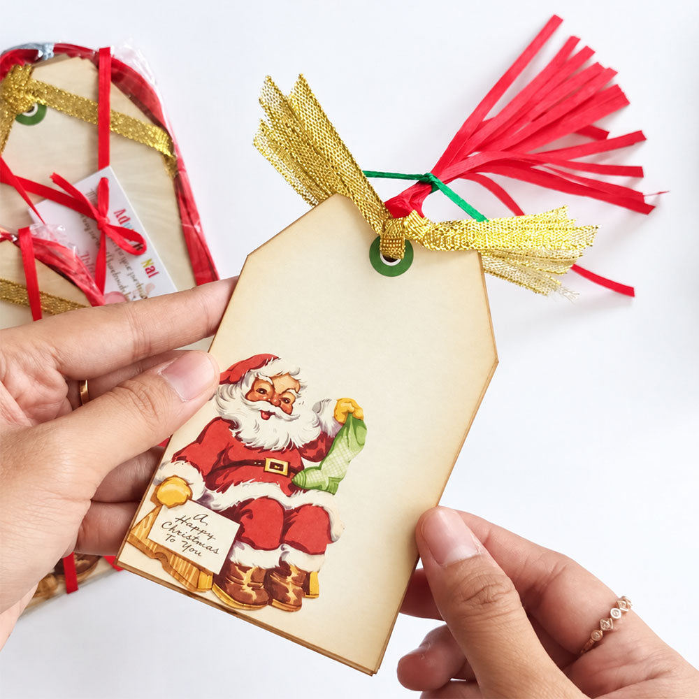Hands holding a vintage Santa gift tag showing the Christmas illustration, greeting, and pre-strung gold ribbon