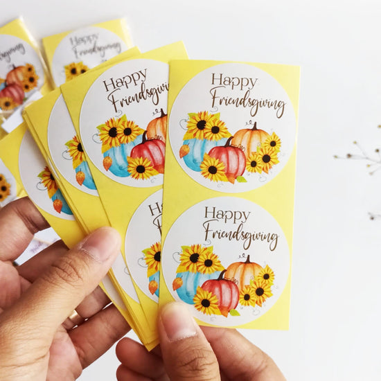 Hand holding set of Happy Friendsgiving stickers with rustic-modern pumpkin and sunflower design for fall favors